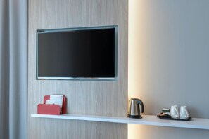 A modern room with a mounted flat-screen TV, a white shelf with a red phone, a kettle, and two white mugs.