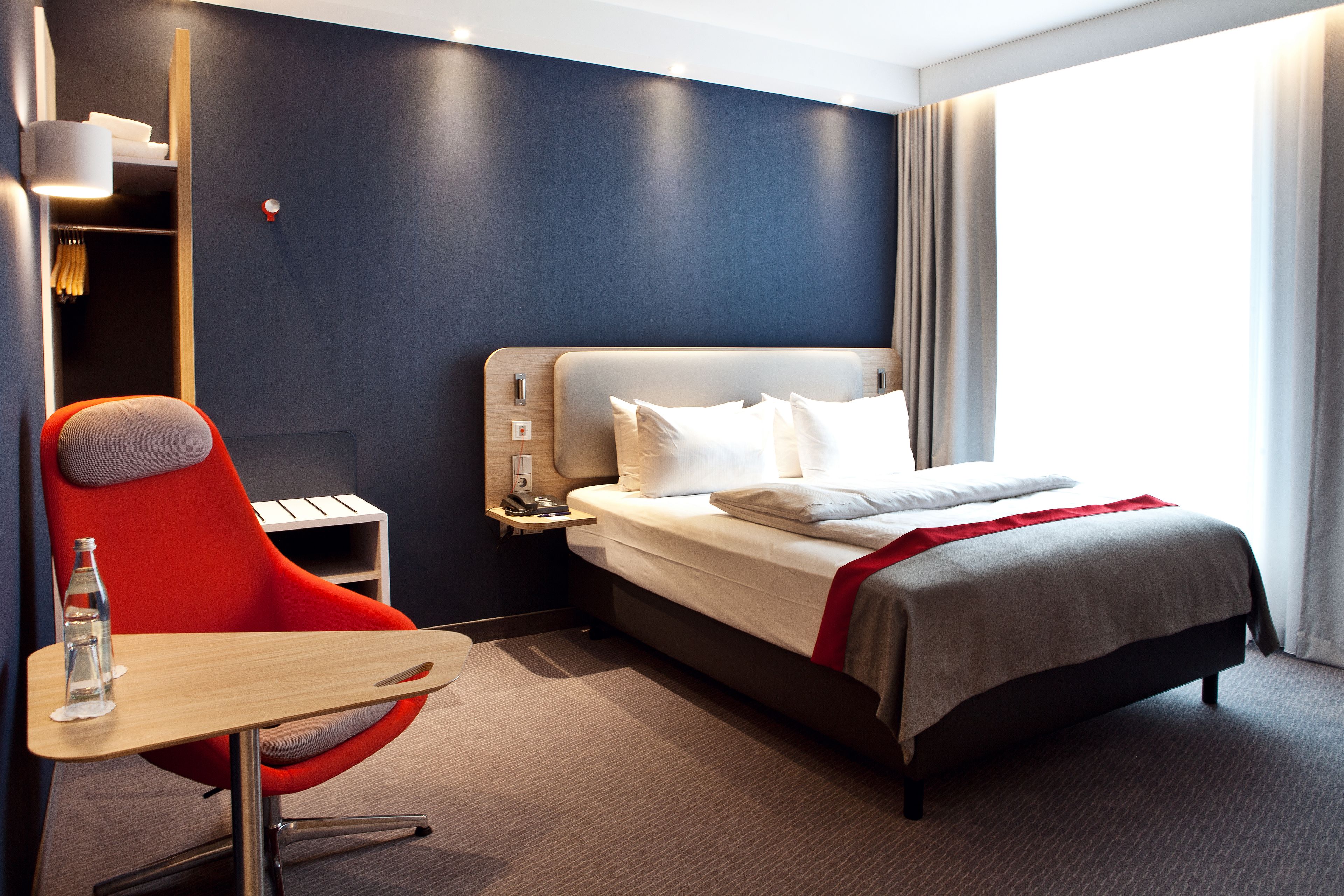 A modern hotel room with a large bed, a red chair, a small table, and a nightstand.