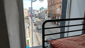 Una habitación con una cama individual, ventana con vista a una calle con edificios y coches estacionados.