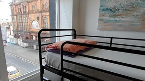 Un dormitorio con una cama litera, una ventana con vistas a un edificio y un cuadro en la pared.