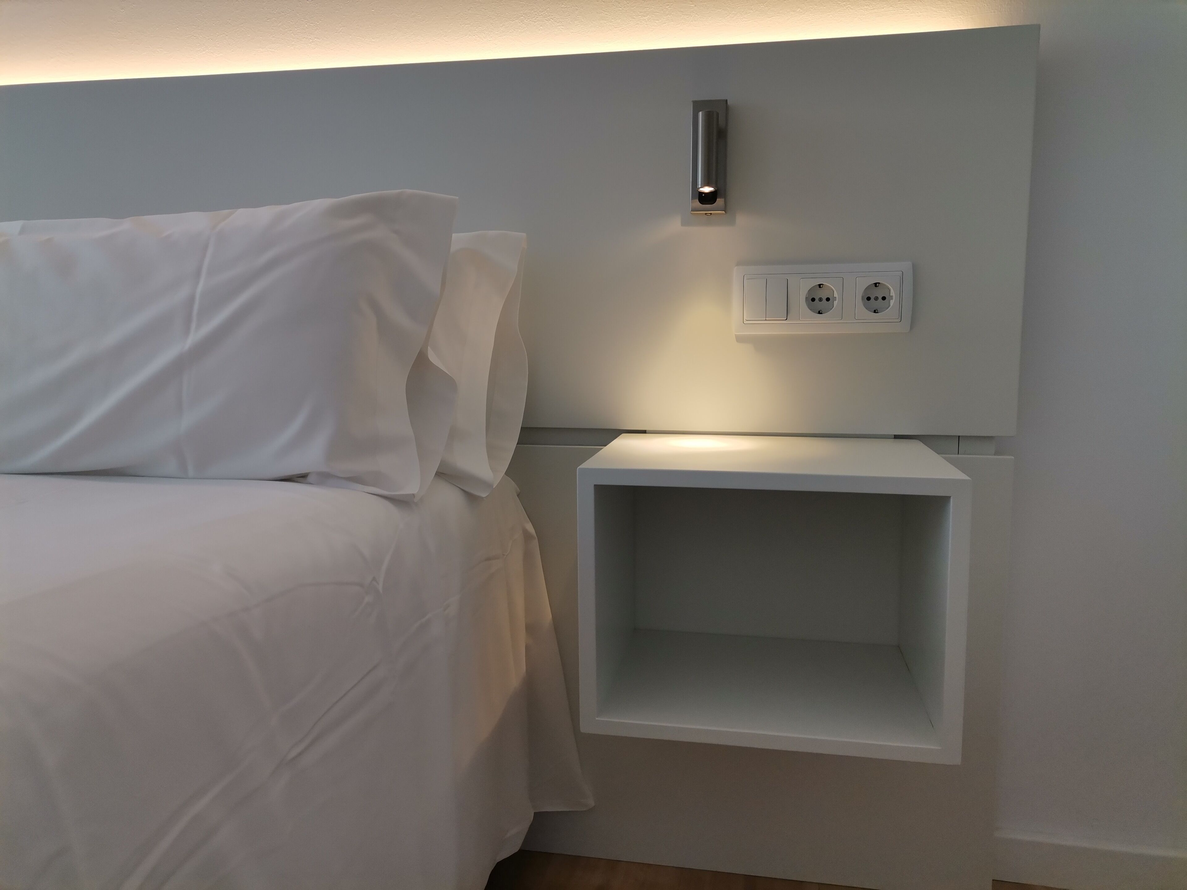 Een net opgemaakt bed met witte lakens, een nachtkastje met een lamp en een stopcontact.