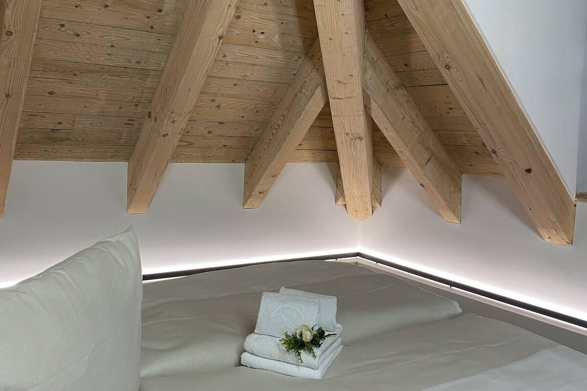 A bedroom with a sloped ceiling supported by wooden beams, a bed with white bedding, and a small decorative item on the bed.