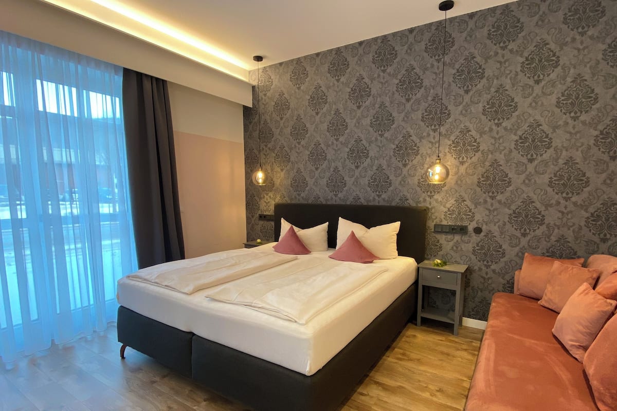 A hotel room with a large bed, two bedside tables, a nightstand, a sofa, and a patterned wallpaper.