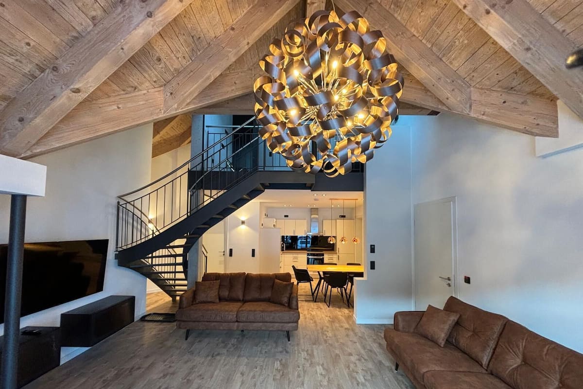A spacious living area with a modern chandelier, a staircase, and a kitchen in the background.