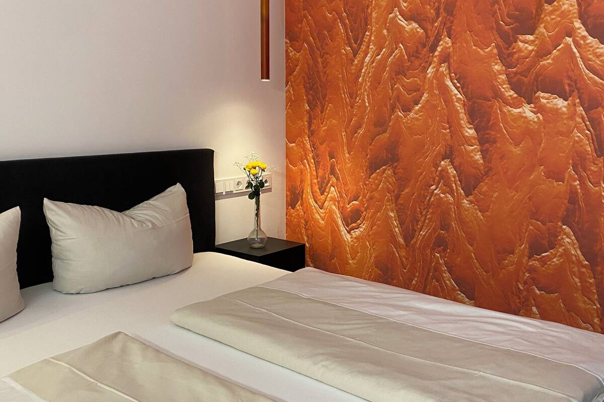 A modern hotel room with a large bed, a black headboard, a nightstand with a vase of flowers, and a textured orange wall.