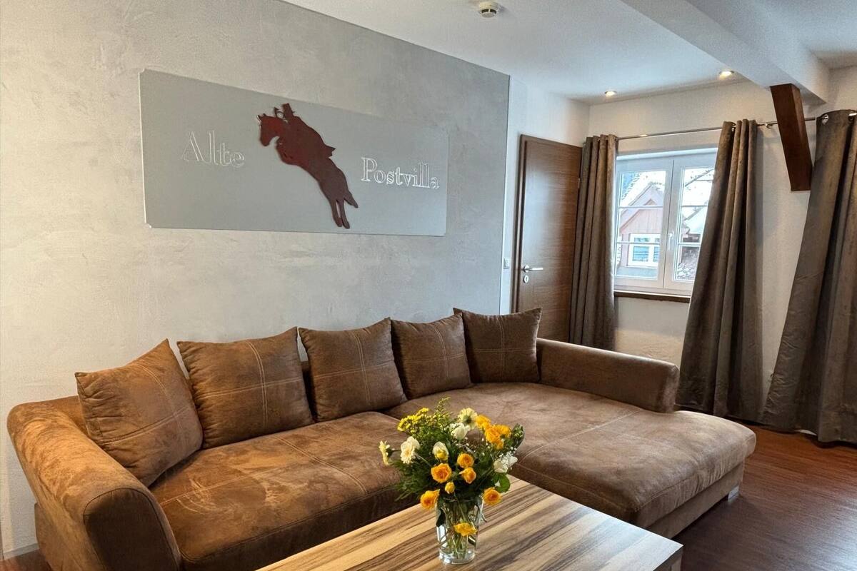 A modern living room with a brown leather sofa, a wooden coffee table with a vase of flowers, and a wall-mounted artwork depicting a horse and the word "Postville".