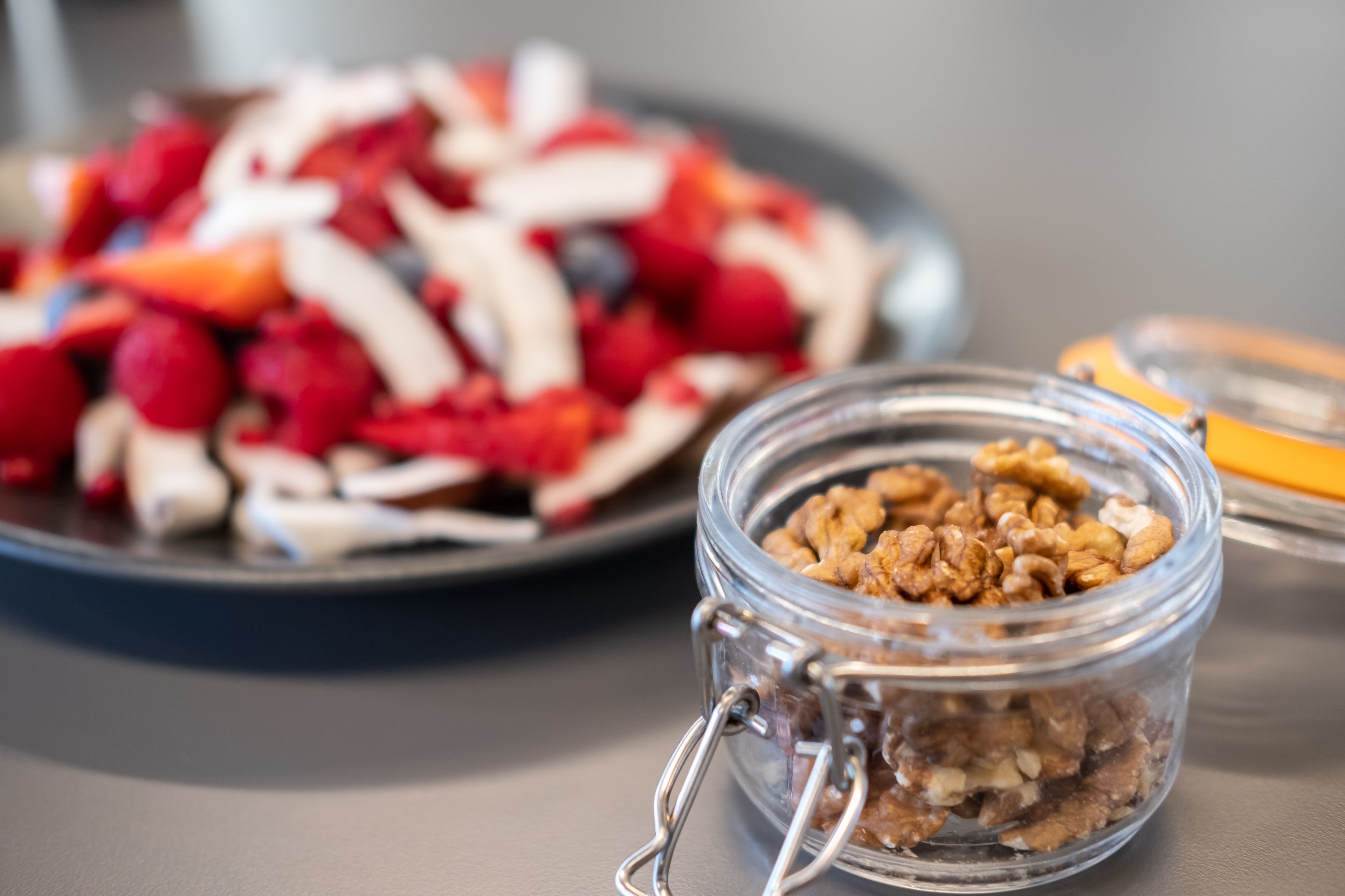Un frasco de vidrio con cierre metálico que contiene nueces, sobre una mesa junto a un plato de frutas y verduras cortadas.