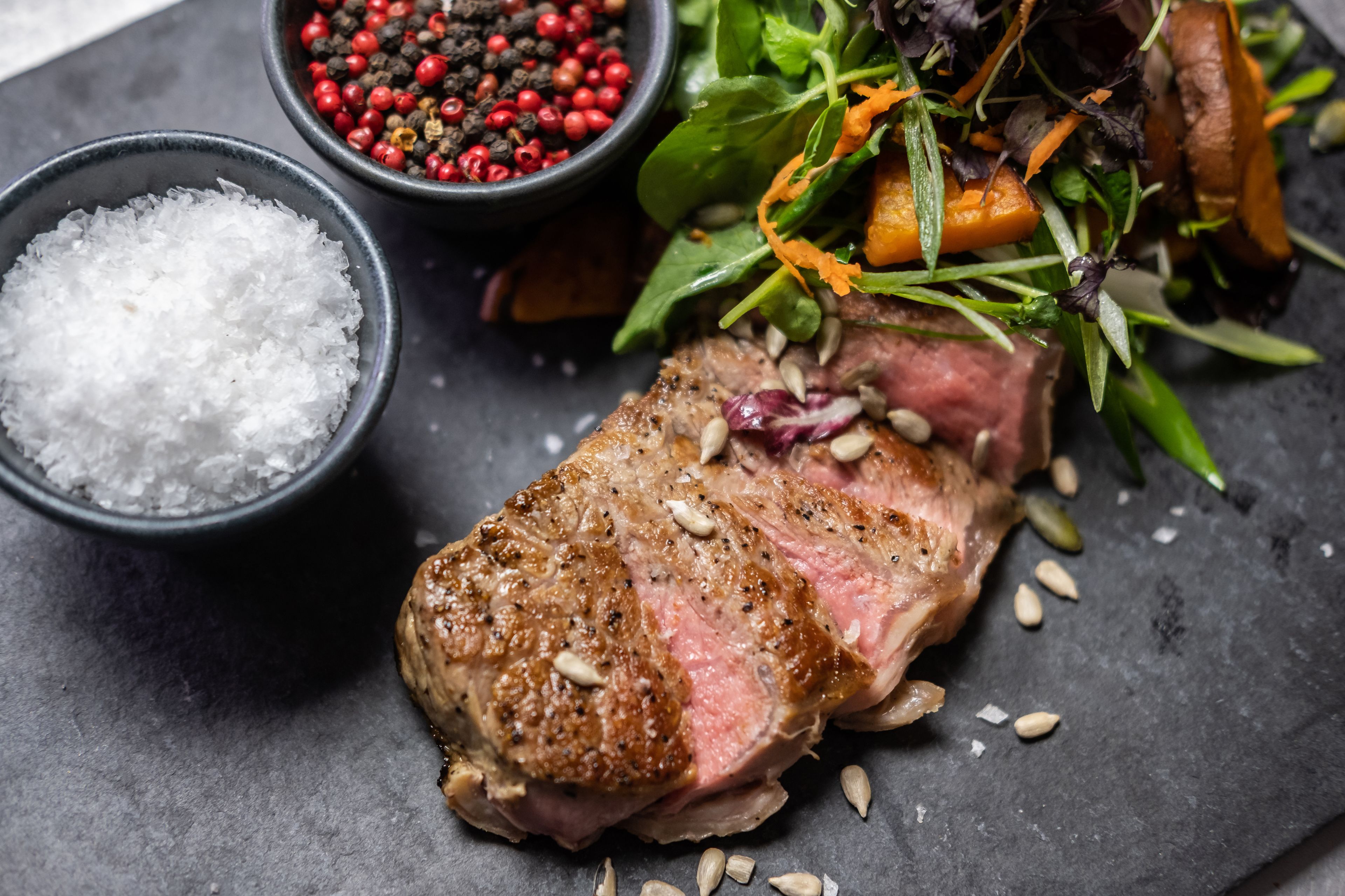 A steak with a side of greens and a bowl of salt.