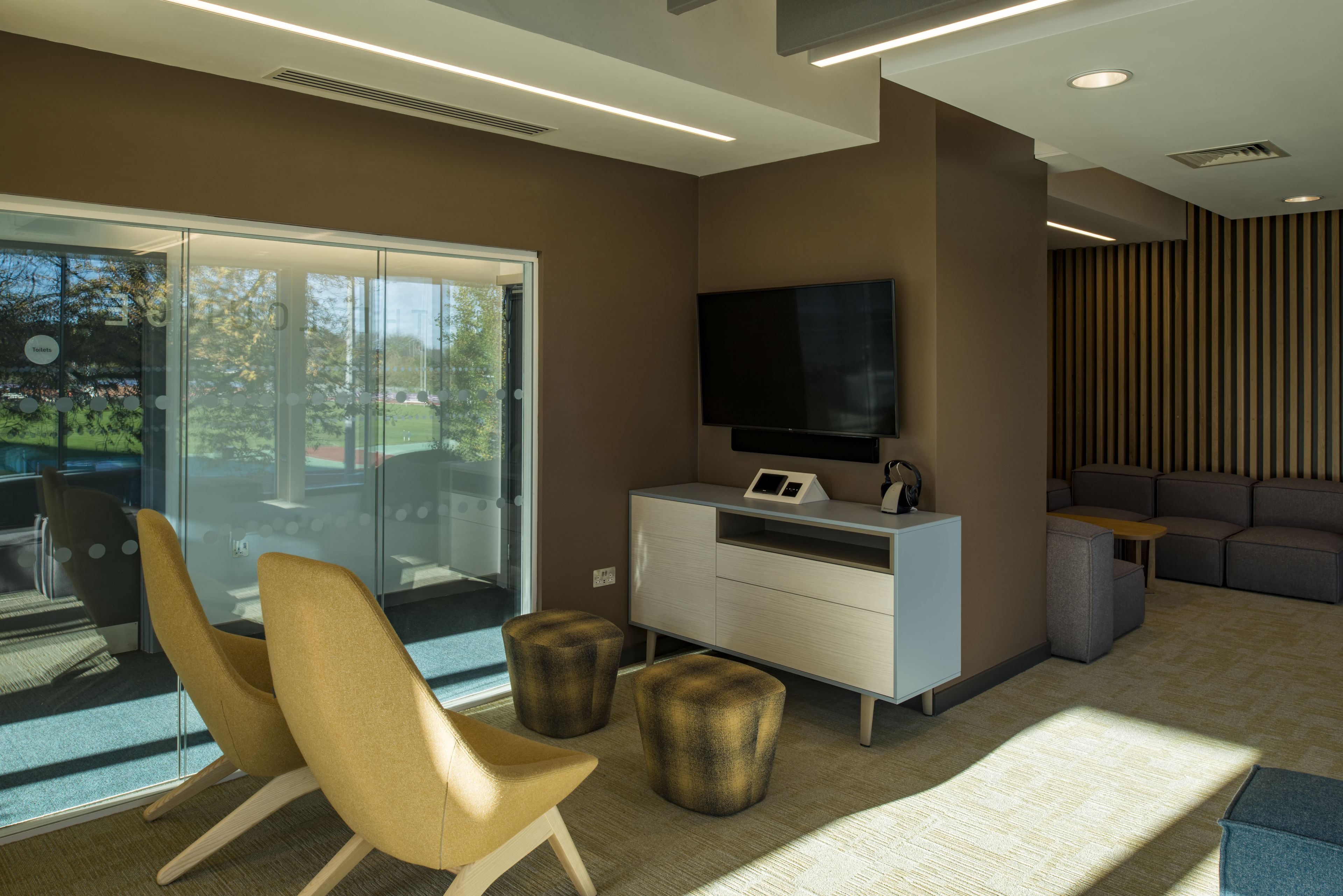 A modern room with a glass door leading outside, a flat-screen TV on a white cabinet, and a seating area with a sofa and ottomans.