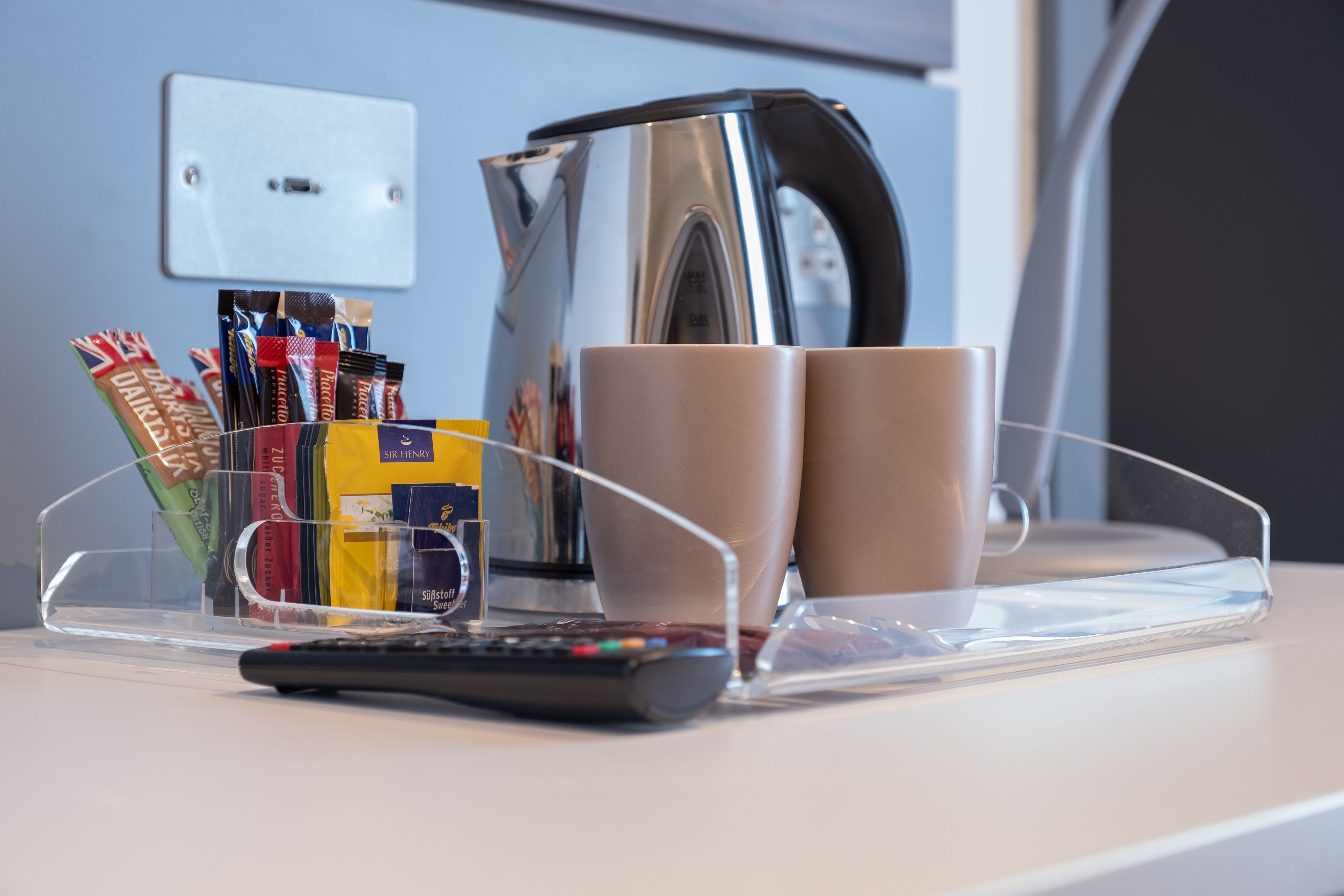 Un plateau en plastique transparent contenant une télécommande, deux tasses et une bouilloire, le tout posé sur une surface blanche.