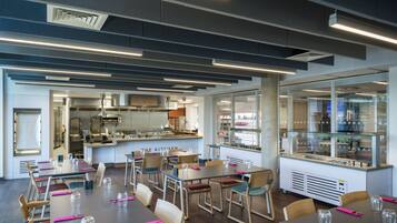 A modern restaurant with multiple dining tables, chairs, and a kitchen area visible through glass partitions.