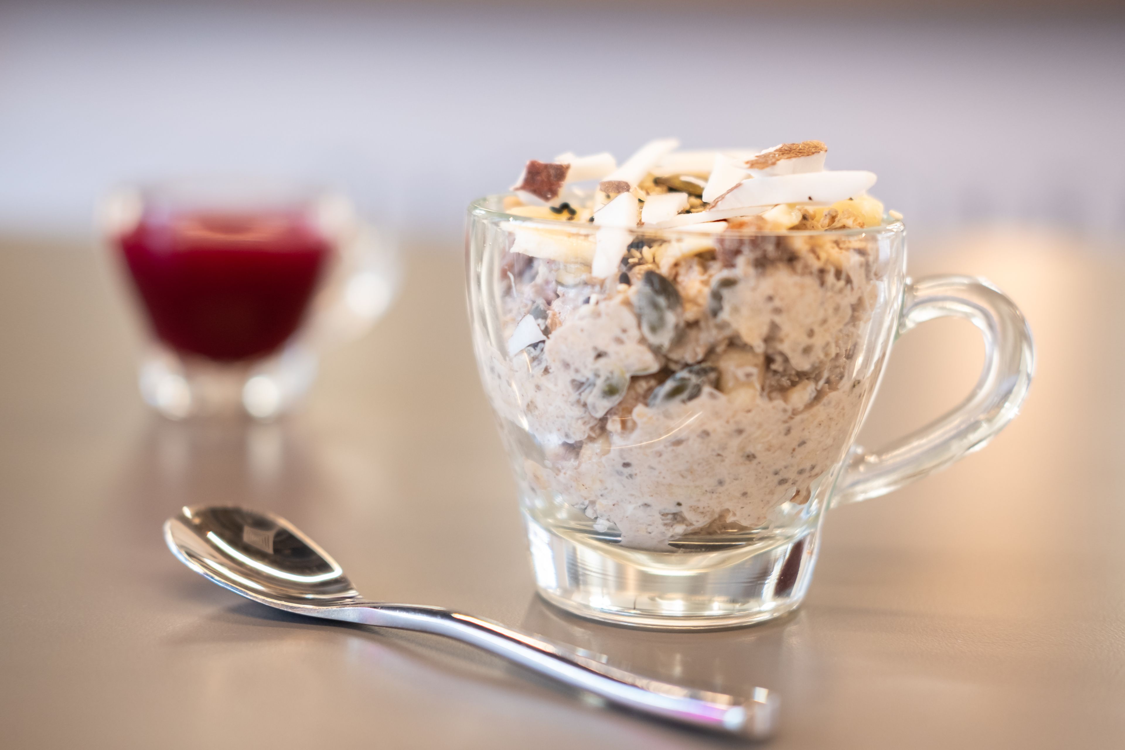Une tasse en verre transparent, avec une cuillère à côté, contenant un dessert à plusieurs couches dans lequel on peut voir des noix et des morceaux de chocolat.