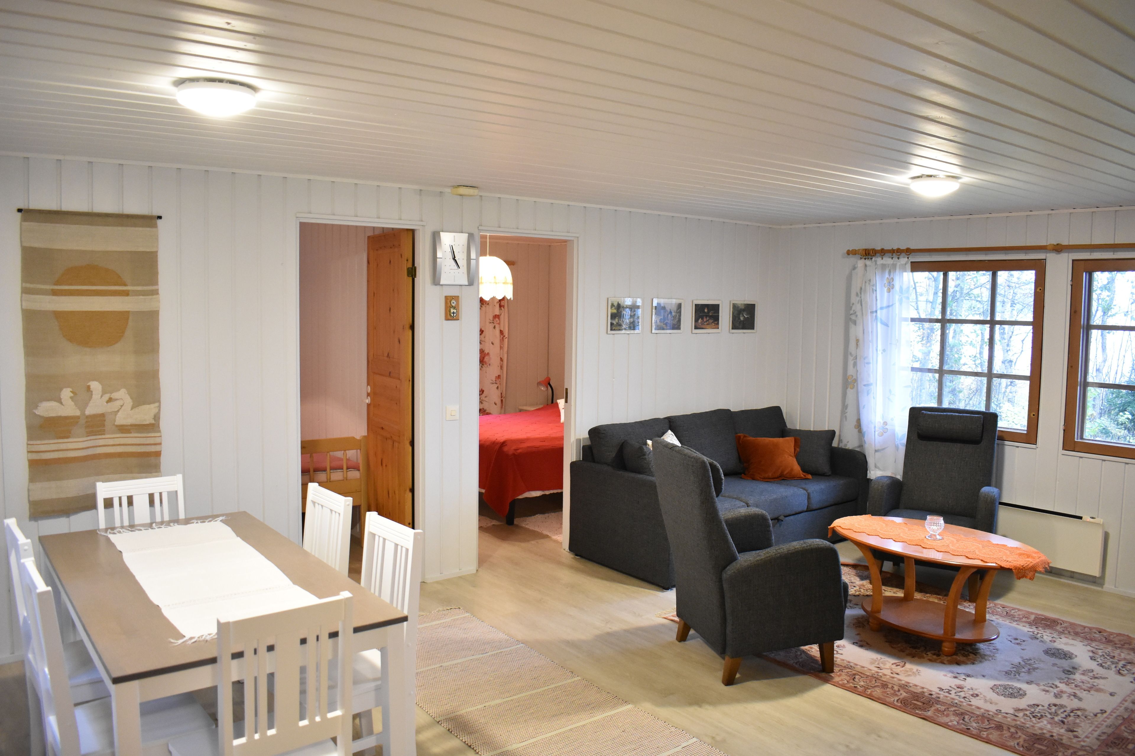 A room with a dining table, chairs, a sofa, and a coffee table. There is a bed in the background and a window with curtains.
