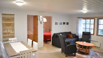 A room with a dining table, chairs, a sofa, and a coffee table. There is a bed in the background and a window with curtains.