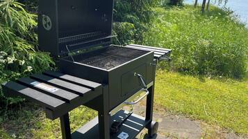 A portable grill with a side table, set on a concrete surface near a grassy area with trees and a body of water in the background.