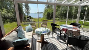 A conservatory with a view of a lake, furnished with chairs and tables, and a wooden bench with cushions. - Karjalan Helmi (Kitee)