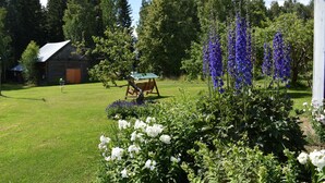 A rustic cabin with a green roof, surrounded by lush greenery and flowering plants. - Karjalan Helmi (Kitee)