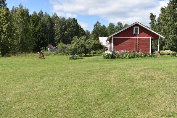 A red barn with a white door and a ladder, surrounded by a grassy field and trees. - Karjalan Helmi (Kitee)