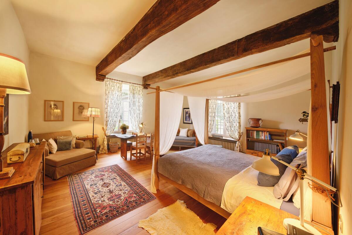 A bedroom with a four-poster bed, a desk with a lamp, a sofa, and a bookshelf.