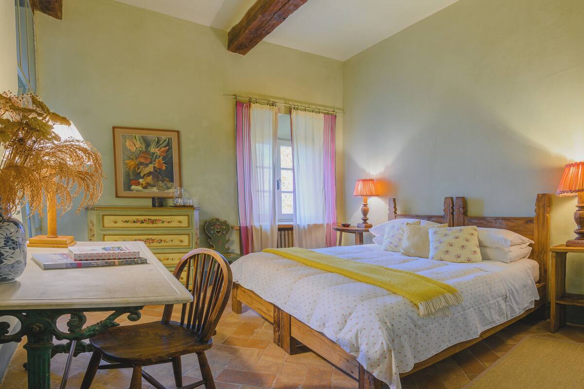 A bedroom with a bed, a chair, a table with books, a lamp, and a window with curtains.