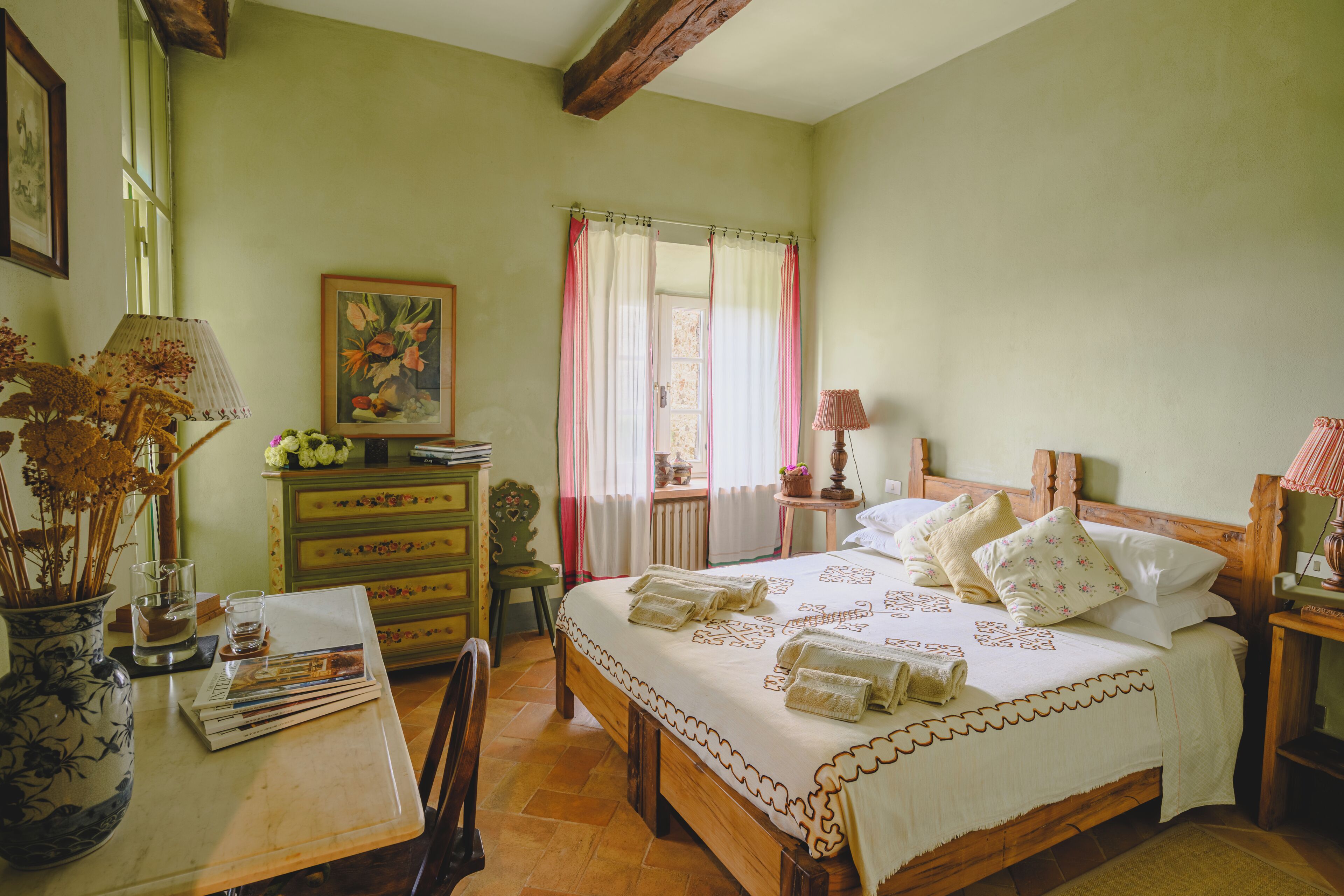 A bedroom with a wooden bed, a bedside table, a chair, a dresser, a window with curtains, and a vase with flowers.