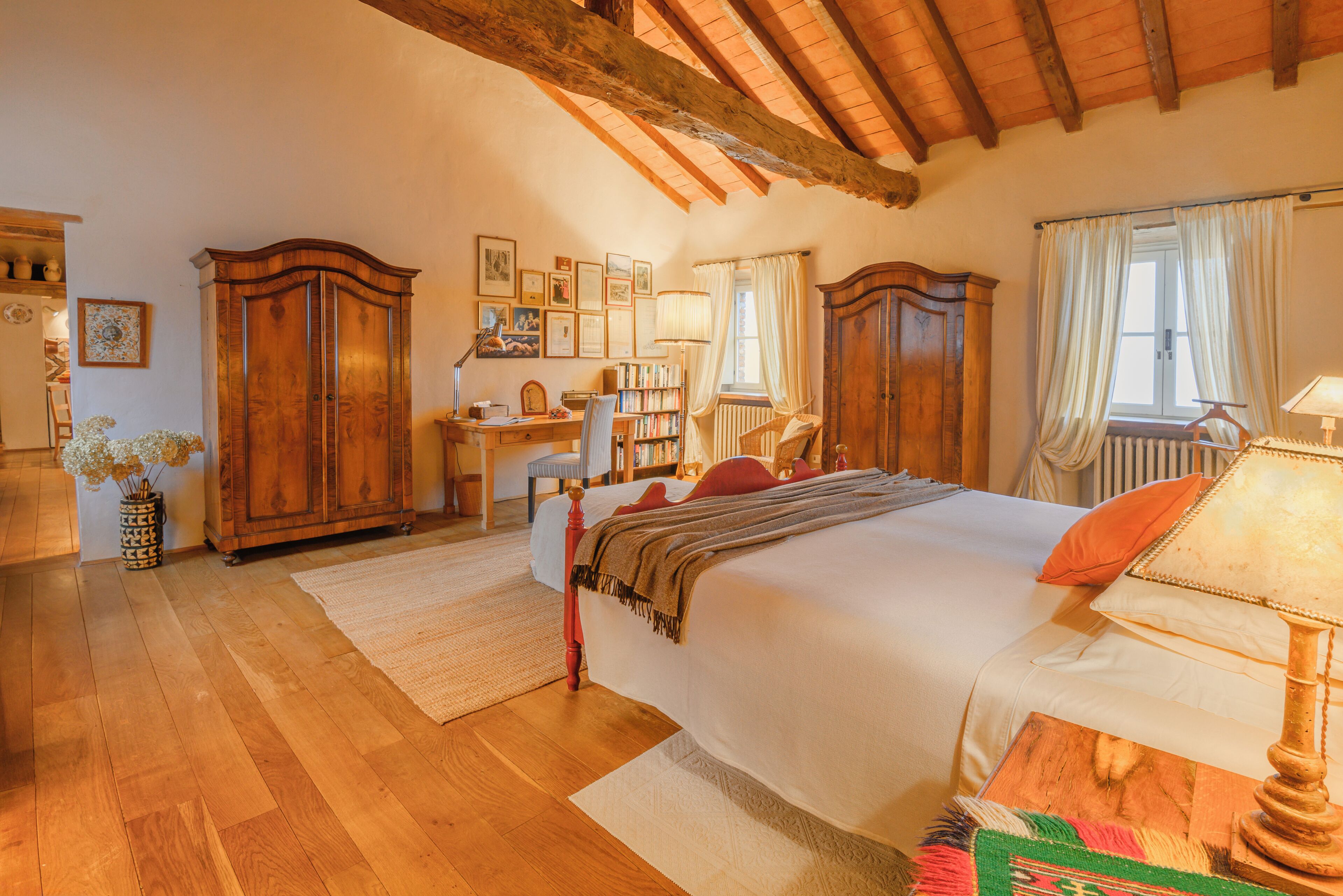 Quarto com teto de madeira, cama com colcha branca, escrivaninha com cadeira e estante de livros.