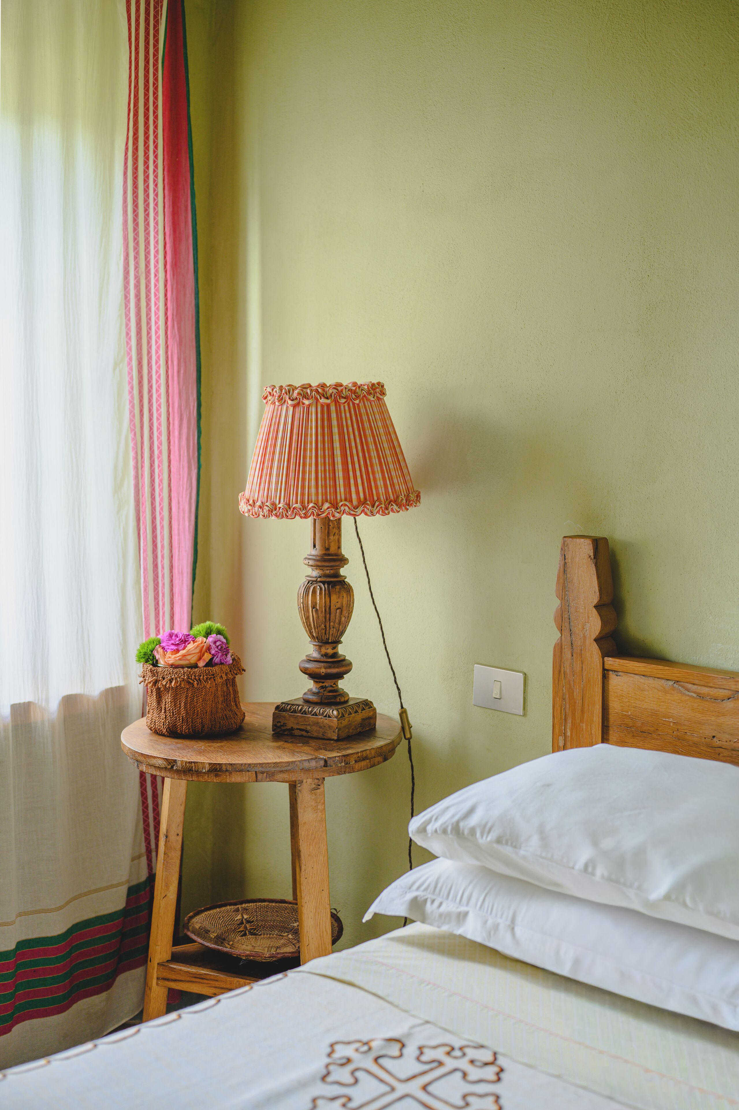 Una cama con almohadas blancas, una mesita de noche de madera con una lámpara, una cesta de mimbre con flores y una cortina a rayas.
