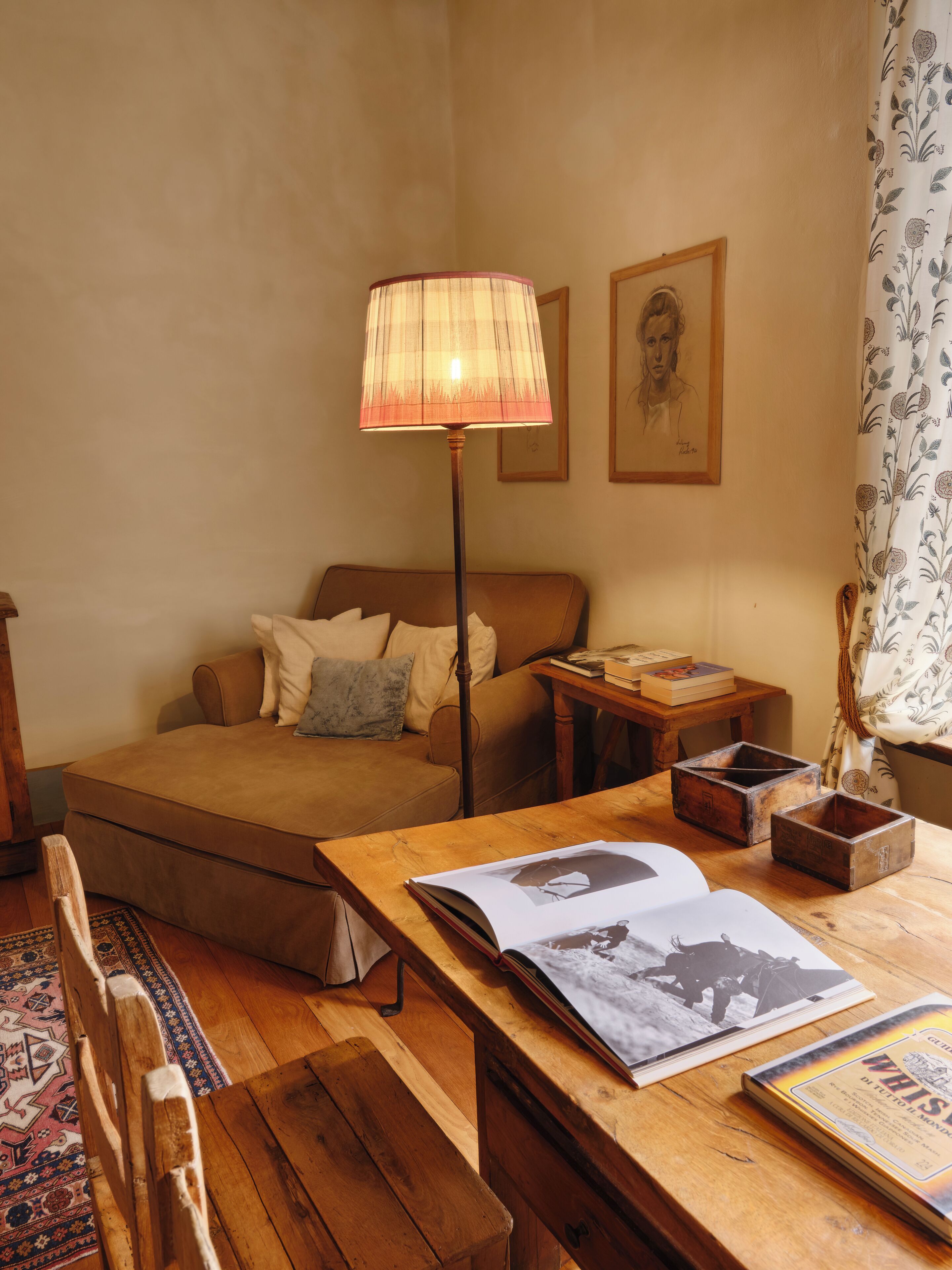 Ein gemütliches Zimmer mit einem Bett, einem Holztisch mit Büchern, einer Lampe und einem gemusterten Vorhang.
