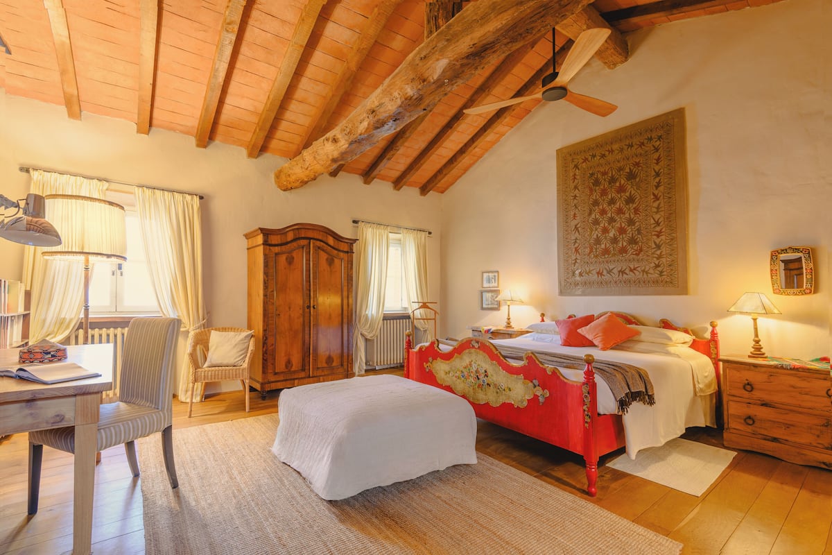 A bedroom with a wooden ceiling, a bed with red and white bedding, a wooden nightstand, a chair, and a desk with a lamp.