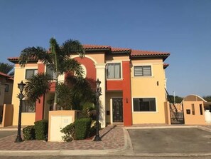 Un edificio residencial de dos plantas con techo de tejas rojas, paredes color beige y un pequeño jardín delantero con palmeras.
