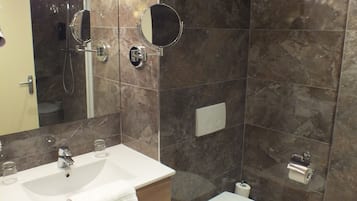 Ein modernes Badezimmer mit einer Marmerdusche, einem Waschbecken inklusive Spiegel, einer Toilette und einem Mülleimer.