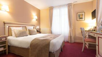 Quarto de hotel com uma cama grande, uma escrivaninha, uma cadeira e uma janela com cortinas.
