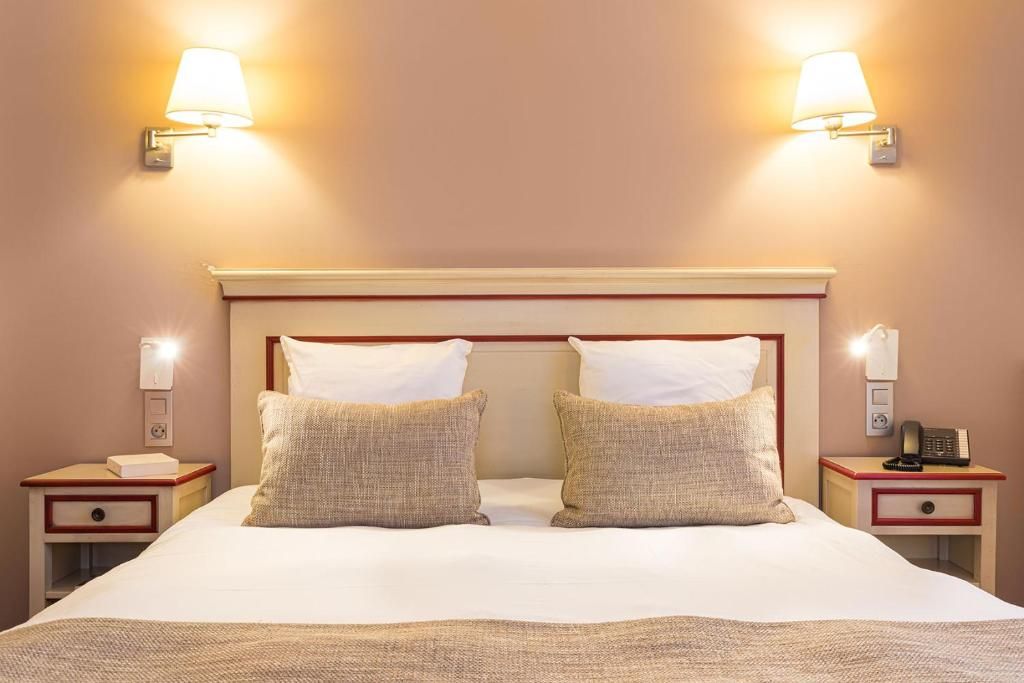 A hotel room with a large bed, two bedside lamps, a nightstand with a phone, and a wall-mounted light fixture.