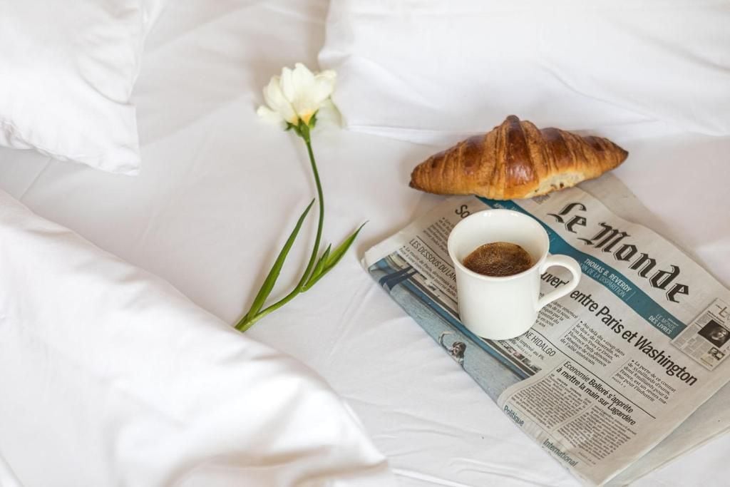 A bed with a white pillow, a white flower, a croissant, a cup of coffee, and a newspaper.
