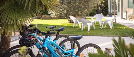 Ein blaues Fahrrad steht in der Nähe einer Palme. Im Hintergrund ist eine Terrasse mit weißen Stühlen und einer Glasveranda zu sehen.