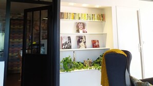Une pièce avec un plafond en bois, une bibliothèque remplie de livres, un fauteuil recouvert d’un plaid jaune et une porte qui donne sur une autre pièce.