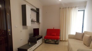 A living room with a TV on a stand, a red sofa, a beige sofa, and a door with a handle. - Amber Apartments (Sarandë)