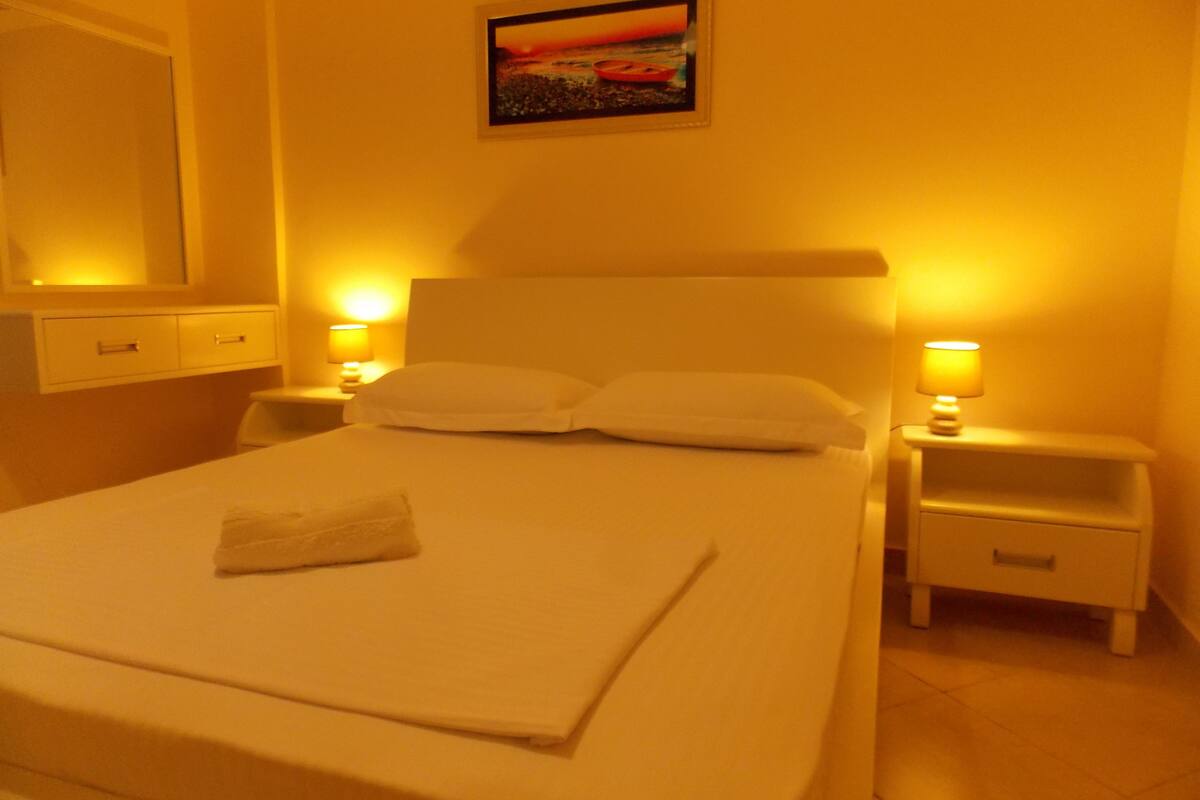 A neatly made bed with a white blanket and a folded towel, two bedside tables with lamps, a nightstand, a painting on the wall, and a mirror above the bed.