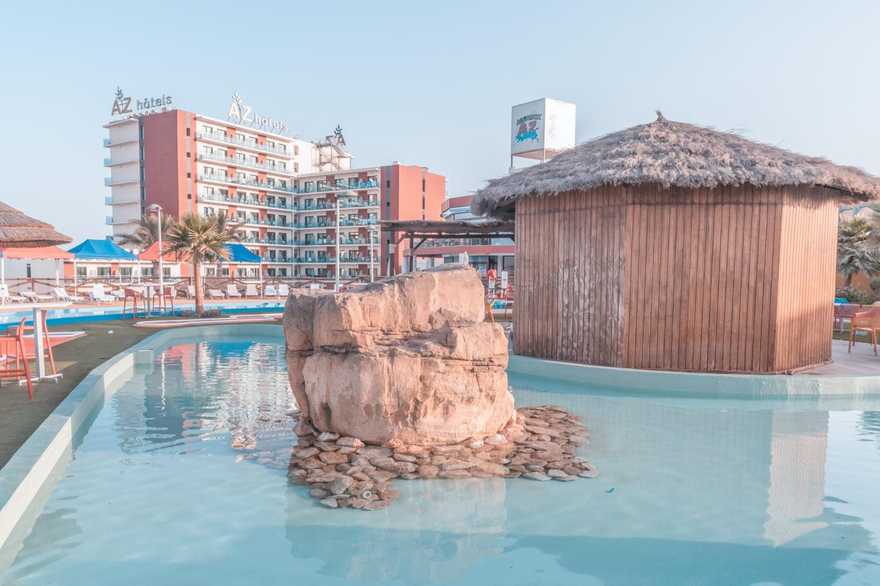 A swimming pool with a rock feature, a wooden hut, and a hotel building in the background.