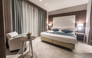 Una habitación de hotel moderna con una cama grande, un área de estar con una silla y una mesa, y vistas de la ciudad.
