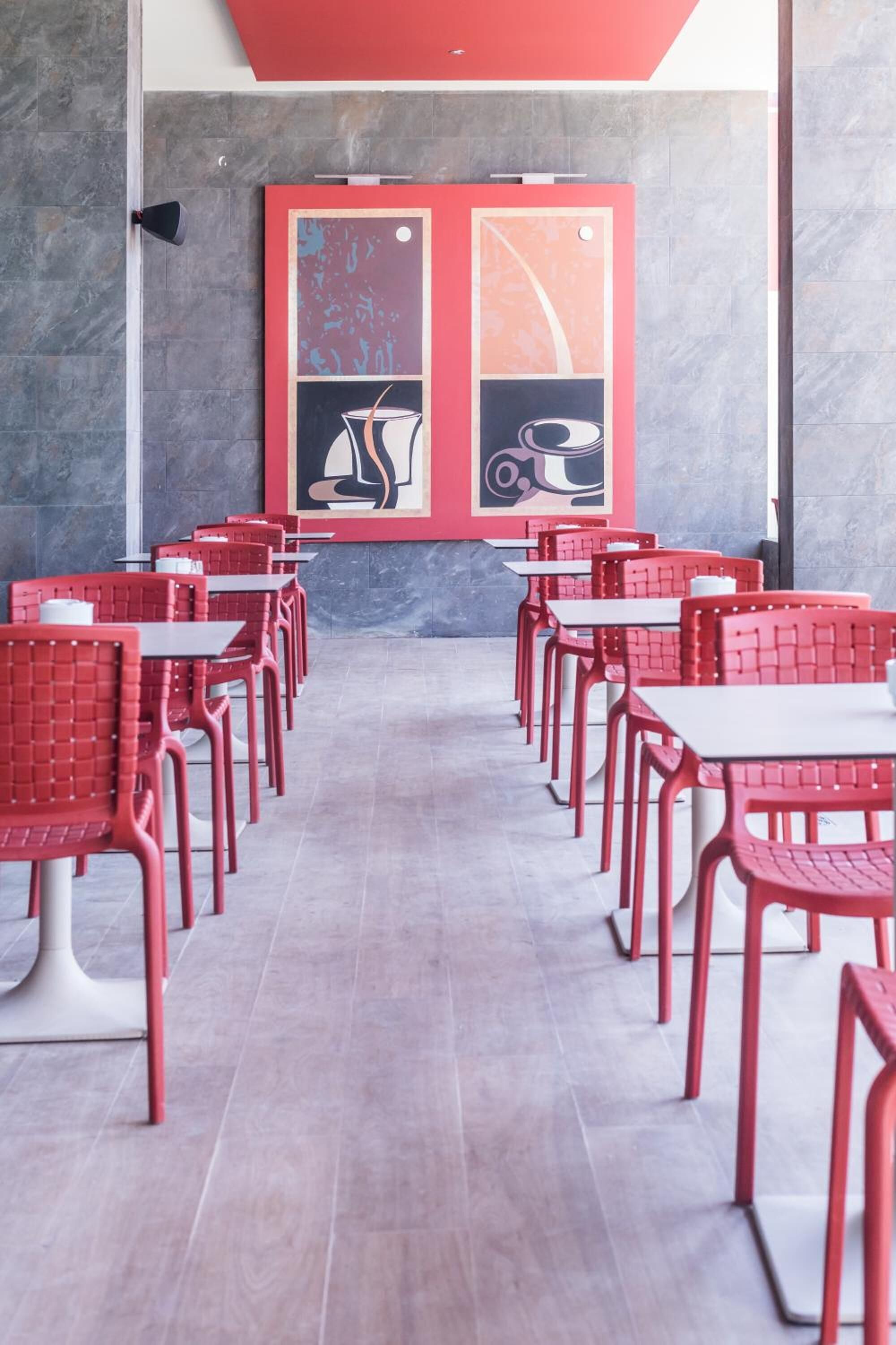 Un café avec des chaises et des tables rouges, un sol carrelé et un mur orné d’œuvres d’art abstraites.