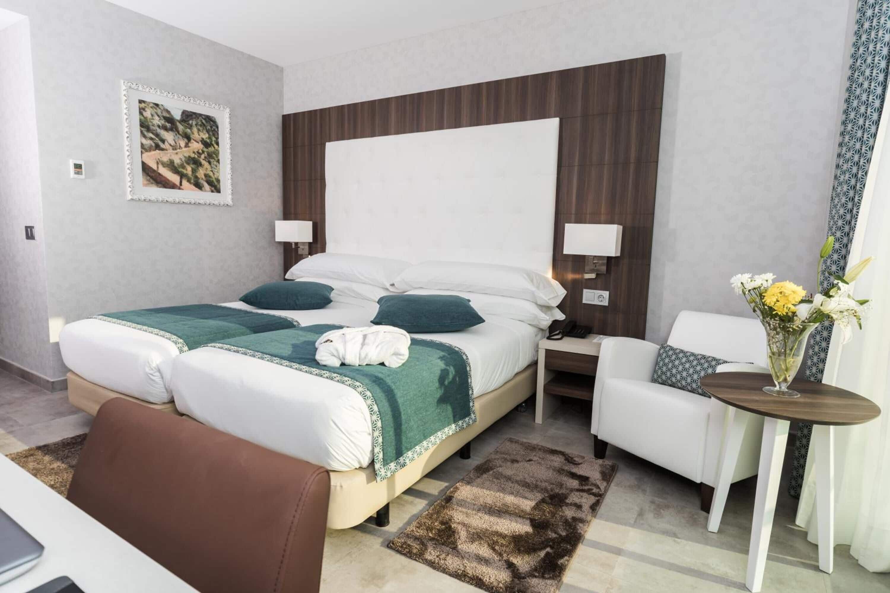 A hotel room with a large bed, bedside tables, a chair, a small table with a vase of flowers, and a framed picture on the wall.