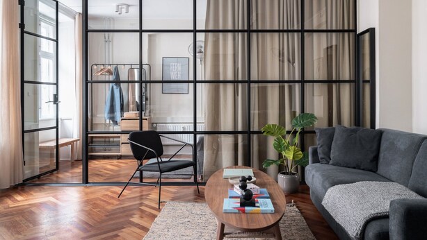A modern living room with a grey sofa, a wooden coffee table, and a black chair. A large window with curtains is visible, and a plant is placed on the floor. - The Brewer apartments by Daniel&Jacob’s (Copenhagen)