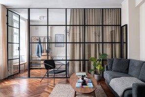 A modern living room with a grey sofa, a wooden coffee table, and a black chair. A large window with curtains is visible, and a plant is placed on the floor. - The Brewers - Daniel&Jacob's Premium Apartments (Copenhagen)