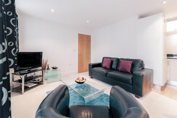 A modern living room with a black leather sofa, a glass coffee table, and a flat-screen TV on a glass stand.