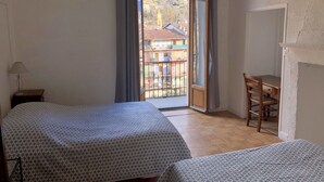 A room with two beds, a desk, and a view of a building through a window. - Hostellerie du Pont Vieux (Sospel)