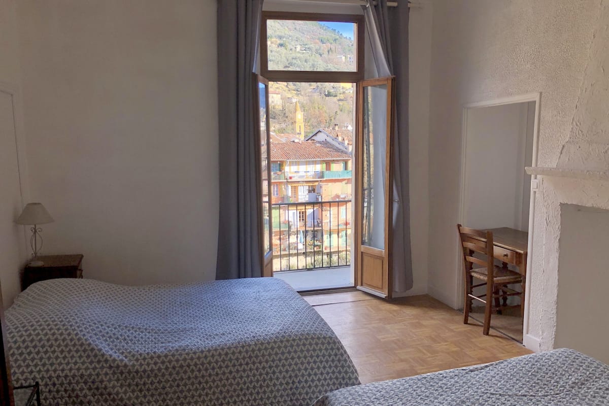 Quarto com duas camas, uma escrivaninha e vista para um edifício através de uma janela.