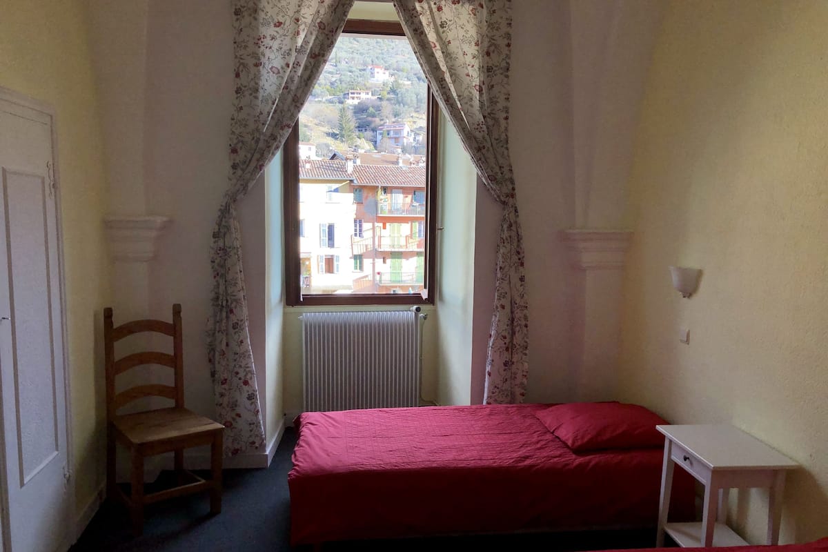 Quarto pequeno com duas camas, uma cadeira, um radiador e uma janela com cortinas.
