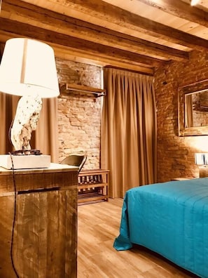 Una habitación con techo de madera, pared de piedra, cama con colcha azul, escritorio con lámpara y una silla.