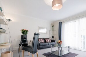 A modern living room with a glass coffee table, a black leather chair, a grey sofa, and a metal shelving unit. - Roomspace Apartments -Cascades Court (London)